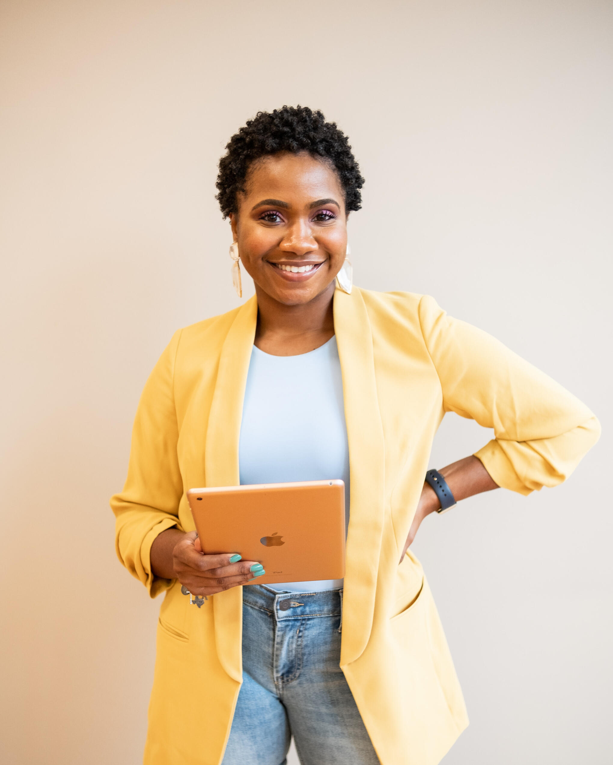 African American woman standing with an Apple iPad in her right hand and her left hand on her hip. She's wearing jeans, a white shirt, and a yellow blazer over top.
