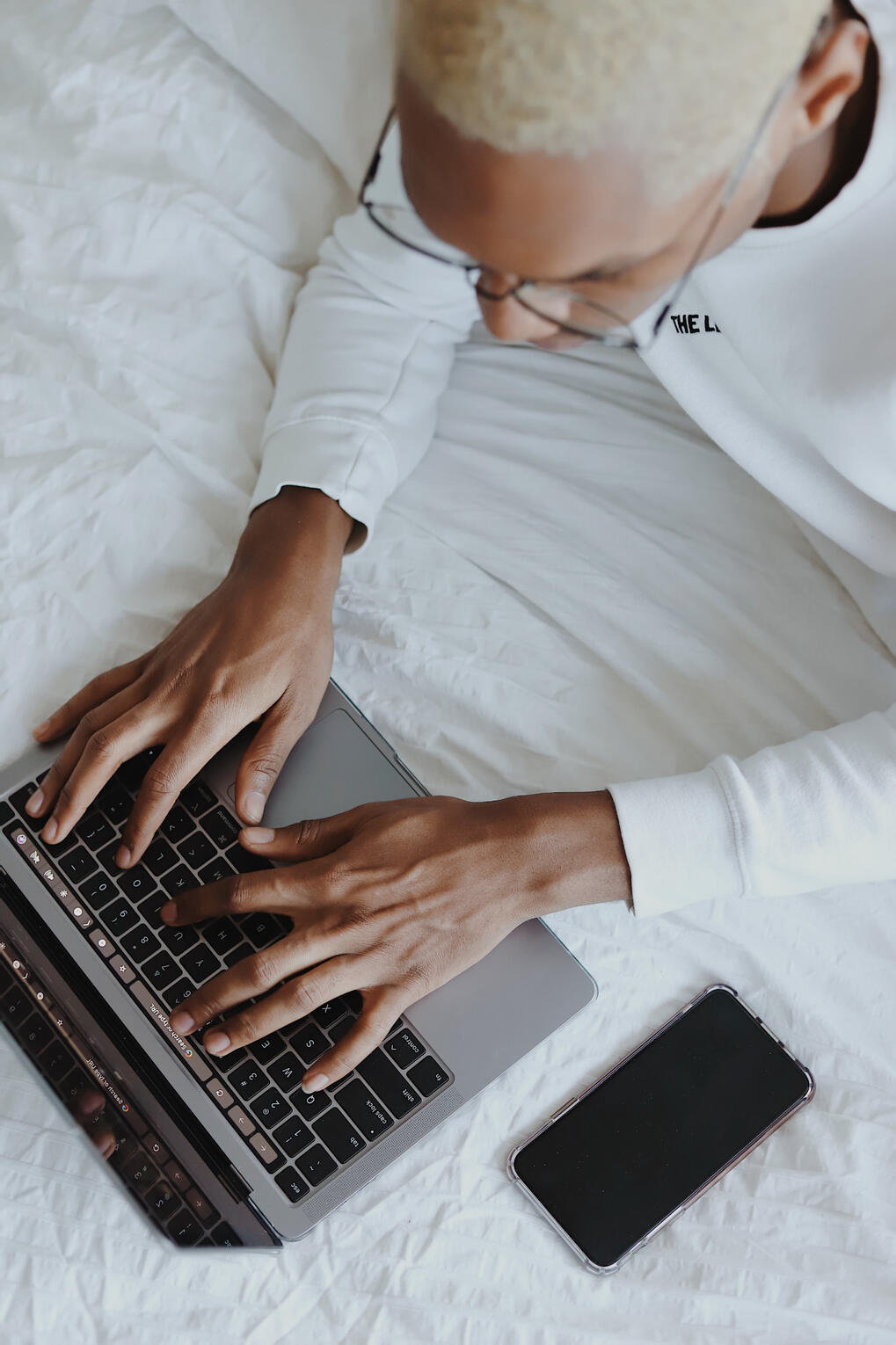 An African American male typing on a new laptop. He's lying on a bed and has blond hair. The bed has a white comforter on top.